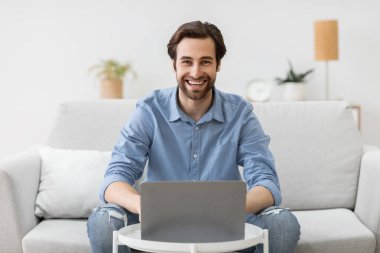 Dizüstü bilgisayardaki adam internette çalışıyor ve evde gülümsüyor.