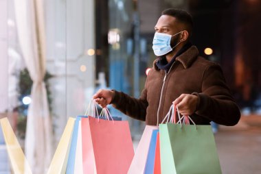 Alışveriş torbalarıyla alışveriş merkezine bakan maskeli siyah bir adam.