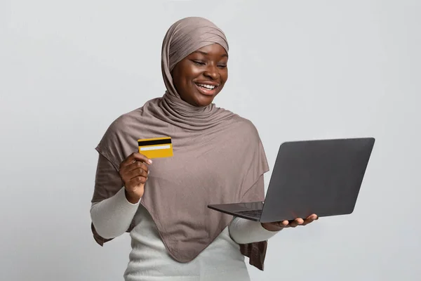 Онлайн оплата. Весела чорна жінка в хіджабі тримає ноутбук і кредитну картку — стокове фото