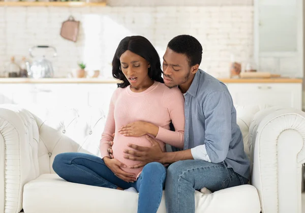 Afroamerykanka w ciąży cierpiąca na ból, mająca skurcze porodowe podczas zamartwiania się mężem pomagającym jej w domu — Zdjęcie stockowe