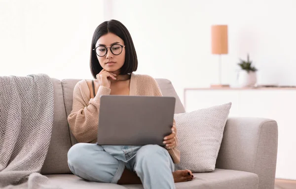 Asian woman sitting on couch, using laptop and thinking — Stock Photo, Image
