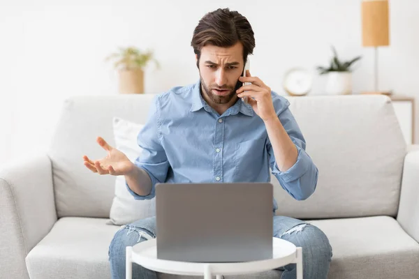 Cep telefonuyla konuşan memnun olmayan adam dizüstü bilgisayarla evde oturuyor. — Stok fotoğraf