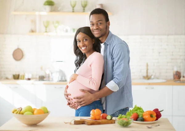 Dieta para un embarazo saludable. Amar a hombre negro abrazando a su hermosa novia embarazada mientras cocina en la cocina —  Fotos de Stock