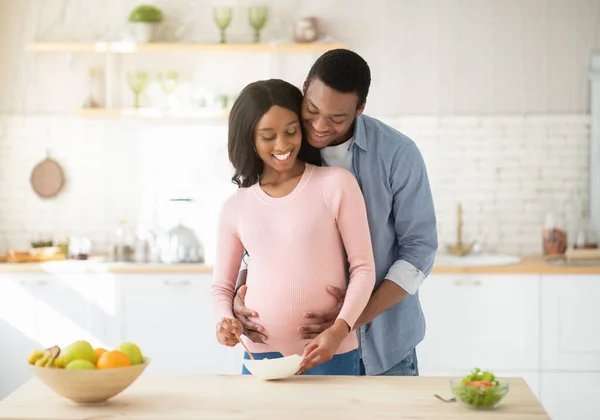 Linda mulher grávida negra fazendo café da manhã saudável na cozinha e marido ajudando-a — Fotografia de Stock