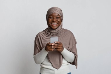 Neşeli siyahi kadın tesettürde. Akıllı telefon tutuyor ve kameraya gülüyor.