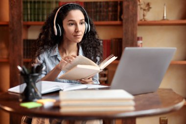Masada oturan, bilgisayar kullanan ve kitap okuyan bir kadın.