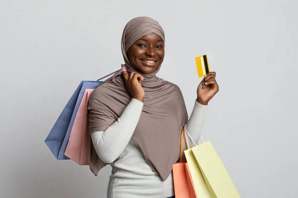 Sonriente mujer negra musulmana en Hijab sosteniendo bolsas de compras y tarjeta de crédito —  Fotos de Stock