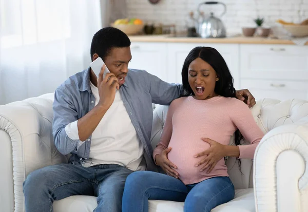 Ожидаемая черная женщина с предродовыми схватками, ее парень звонит доктору по смартфону из дома — стоковое фото