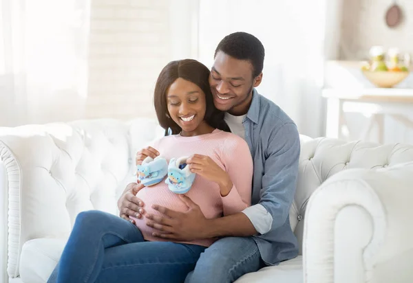 Портрет позитивної вагітної пари з крихітними дитячими пінетками, що обіймаються на дивані в приміщенні — стокове фото