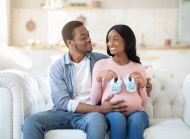 Mutlu siyah çift bebek bekliyor, kanepede birlikte oturuyorlar ve ellerinde küçük çocuk ayakkabıları var.