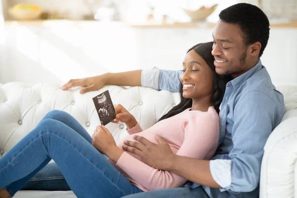 Portrait of positive black couple with ultrasound photo of their future baby cuddling on sofa at home — Stok Foto