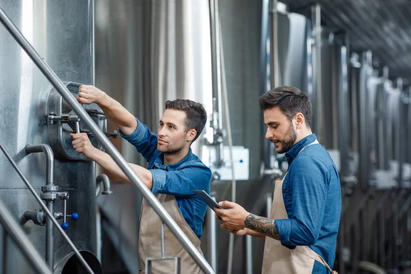 Millennial chico trabajador en delantal gira una válvula en la caldera grande para la cerveza, gerente con el proceso de control de tabletas en el interior — Foto de Stock