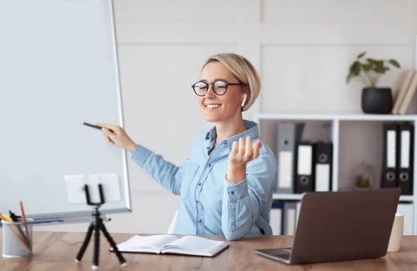 Profesor milenario positivo apuntando a pizarra vacía cerca de la computadora portátil en casa, dando conferencia en línea, espacio para el diseño —  Fotos de Stock