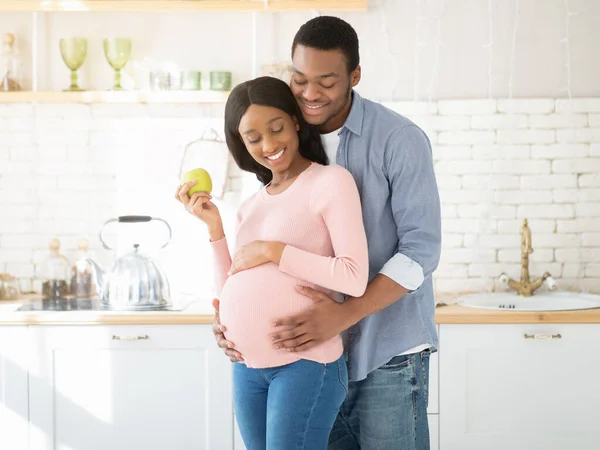Retrato de cara negro amoroso abraçando sua linda esposa expectante com maçã na cozinha — Fotografia de Stock