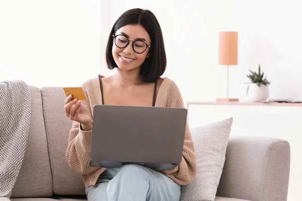 Asian lady in glasses making purchases using laptop holding card — Stock Photo, Image