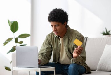 Genç siyah adam evdeki dizüstü bilgisayarında online alışveriş için kredi kartı kullanıyor.
