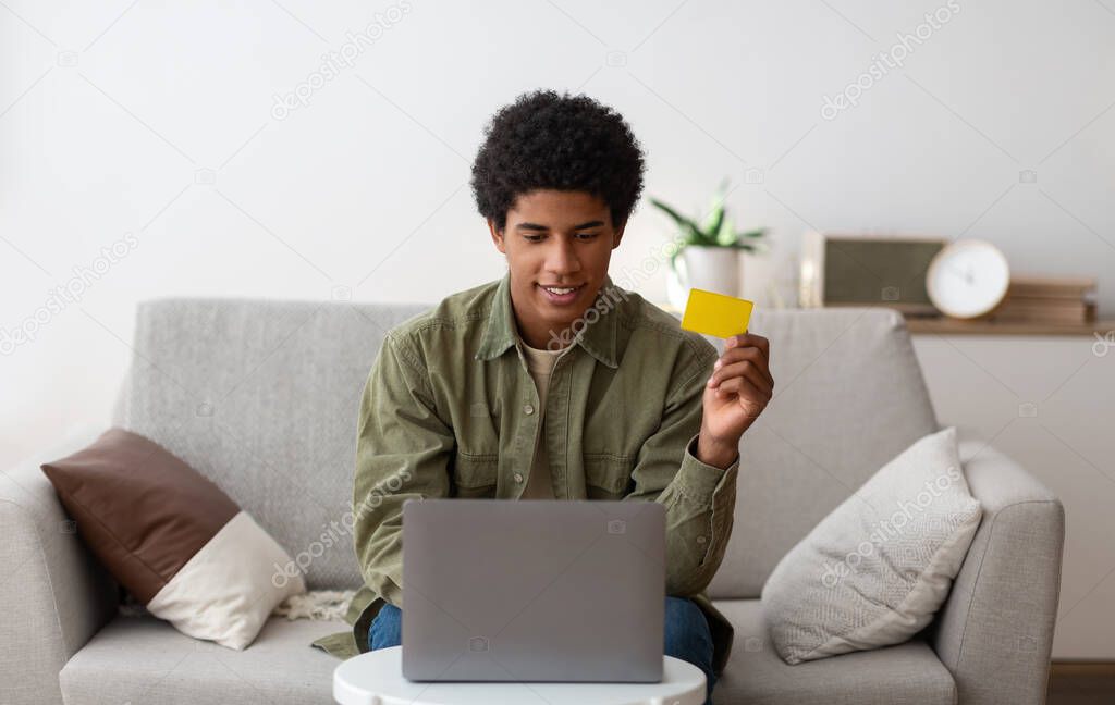 Online shopping. Portrait of cheerful black teenager using credit card and laptop for purchasing goods on web from home