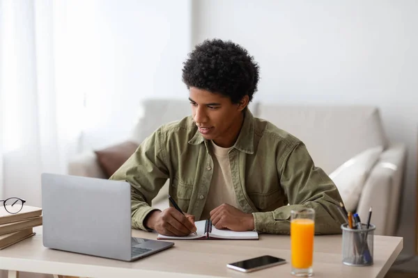 Concepto de educación en el hogar. Estudiante negro escribiendo en copybook durante conferencia en línea o lección web en casa — Foto de Stock