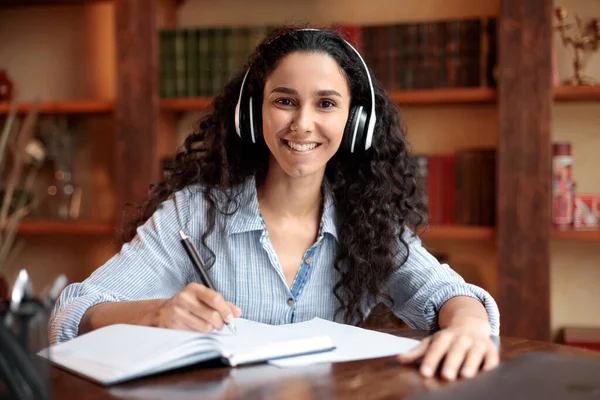 Frau sitzt am Schreibtisch, trägt Headset und schreibt in Notizbuch — Stockfoto