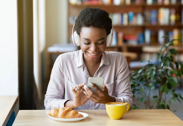 Jovencita alegre negra en auriculares eligiendo la lista de reproducción de música en su teléfono inteligente en la cafetería urbana —  Fotos de Stock