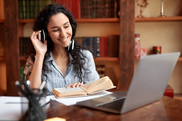 Dame sitzt am Schreibtisch, berührt Headset, benutzt Computer, liest Buch — Stockfoto