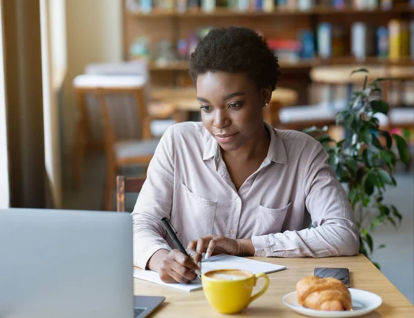 Millennial donna di colore che studia o lavora online, prendendo appunti in copybook al caffè urbano — Foto Stock