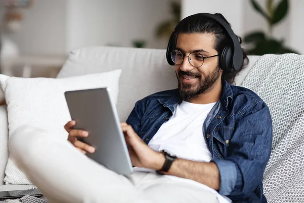Guapo indio en auriculares inalámbricos relajante con tableta digital en casa —  Fotos de Stock