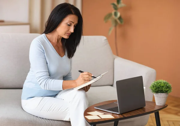 Mature woman writing and using laptop at home — Stock Photo, Image