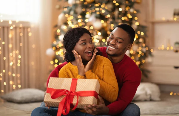 Surpreso milenar afro-americano mulher olha para o marido, ele dá presente com fita vermelha na sala de estar — Fotografia de Stock