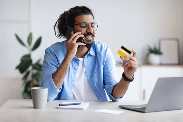 Pagamentos fáceis. Indiano Freelancer Guy Segurando cartão de crédito e falando no celular — Fotografia de Stock