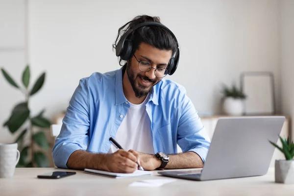 Western Guy φορώντας ακουστικά που μελετούν σε απευθείας σύνδεση με το φορητό υπολογιστή στο σπίτι, λαμβάνοντας σημειώσεις — Φωτογραφία Αρχείου