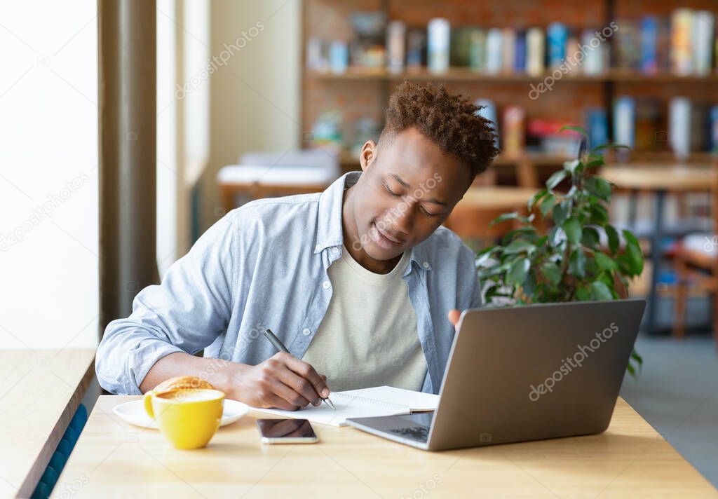 Positive black man using laptop for online work or studies, writing in notebook, doing project at cozy city cafe