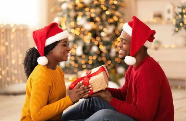 Šťastný mladý africký Američan muž a žena v Santa klobouky výměna dárků v interiéru — Stock fotografie