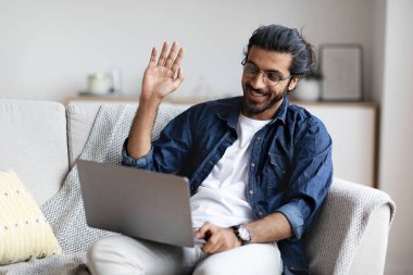 Neşeli genç Hintli adam evdeki bilgisayarında görüntülü arama yapıyor.