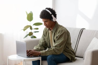 Black teen guy watching educational webinar on laptop, wearing headphones, taking part in web lesson at home