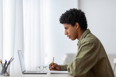 Side view of millennial black student sitting at desk with laptop, making notes in copybook, learning remotely from home