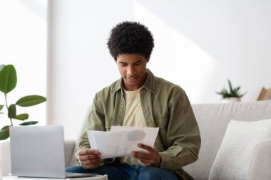 Happy black guy learning online from home, writing coursework, holding documents near laptop