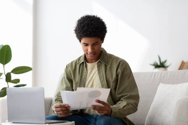 Happy black guy learning online from home, writing coursework, holding documents near laptop — Fotografia de Stock
