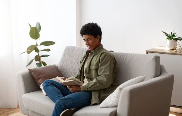 Cheerful black male student sitting on couch with book, reading and enjoying home studies — Foto de Stock