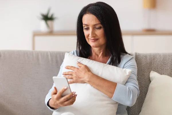 Smiling mature woman using mobile phone, holding pillow — Photo