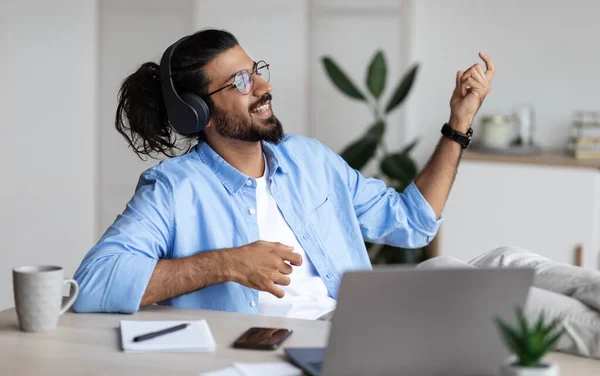 Přestávka v práci. Radostný africký podnikatel poslouchající hudbu ve sluchátkách a hrající na virtuální kytaru, relaxující na pracovišti v kanceláři. — Stock fotografie