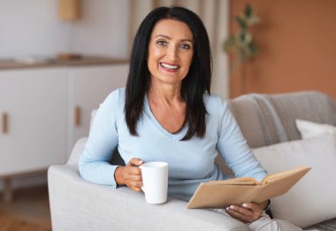 Kitap okuyan ve elinde kahve tutan olgun bir kadın.