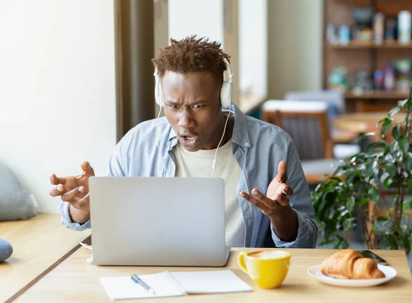 Stressed African American guy in headphones using laptop to work on complicated business project, facing problem at cafe —  Fotos de Stock