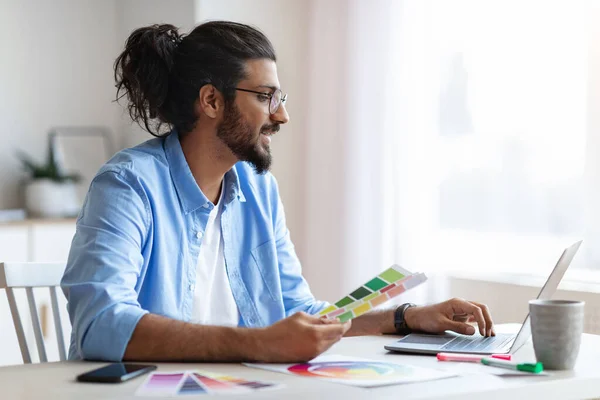 Ung västerländsk inredningsarkitekt som håller färgprover och arbetar på laptop — Stockfoto