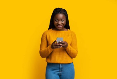 Smiling African American Lady Holding Smartphone, Texting With Friends On Yellow Background