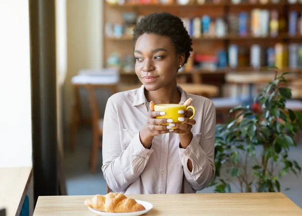 Pretty African American woman having breakfast, enjoying her morning coffee with croissant at city cafe — Stok fotoğraf