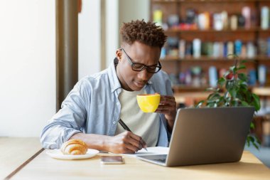 Havalı siyahi adamın kafede dizüstü bilgisayar dersi alması, ders sırasında not alması, sıcak kahve içmesi.
