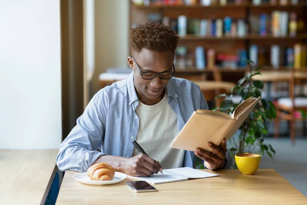 Funky ragazzo nero con libro di testo prendere appunti in copybook in accogliente caffè — Foto Stock