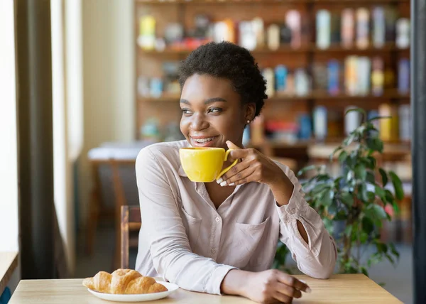 Happy black lady with cup of coffee and croissant enjoying peaceful morning, looking out window at cozy urban cafe