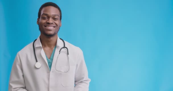 Alegre médico profesional afroamericano mostrando espacio vacío para maqueta, señalando a un lado un fondo de estudio azul — Vídeos de Stock